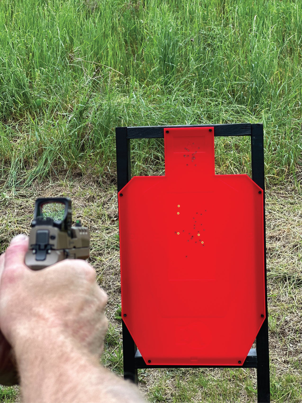 A person aims a handgun at a red target with multiple holes, set in a grassy outdoor area.